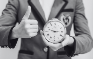 A man in a suit holding up a clock and giving a thumbs up.