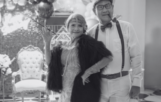 An older couple dressed in 1920s costume posing beside an armchair.