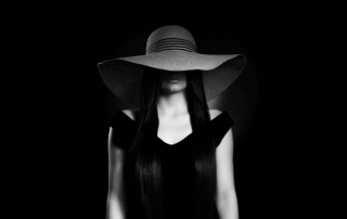 A woman in a wide-brimmed hat with her face hidden, against a black background.