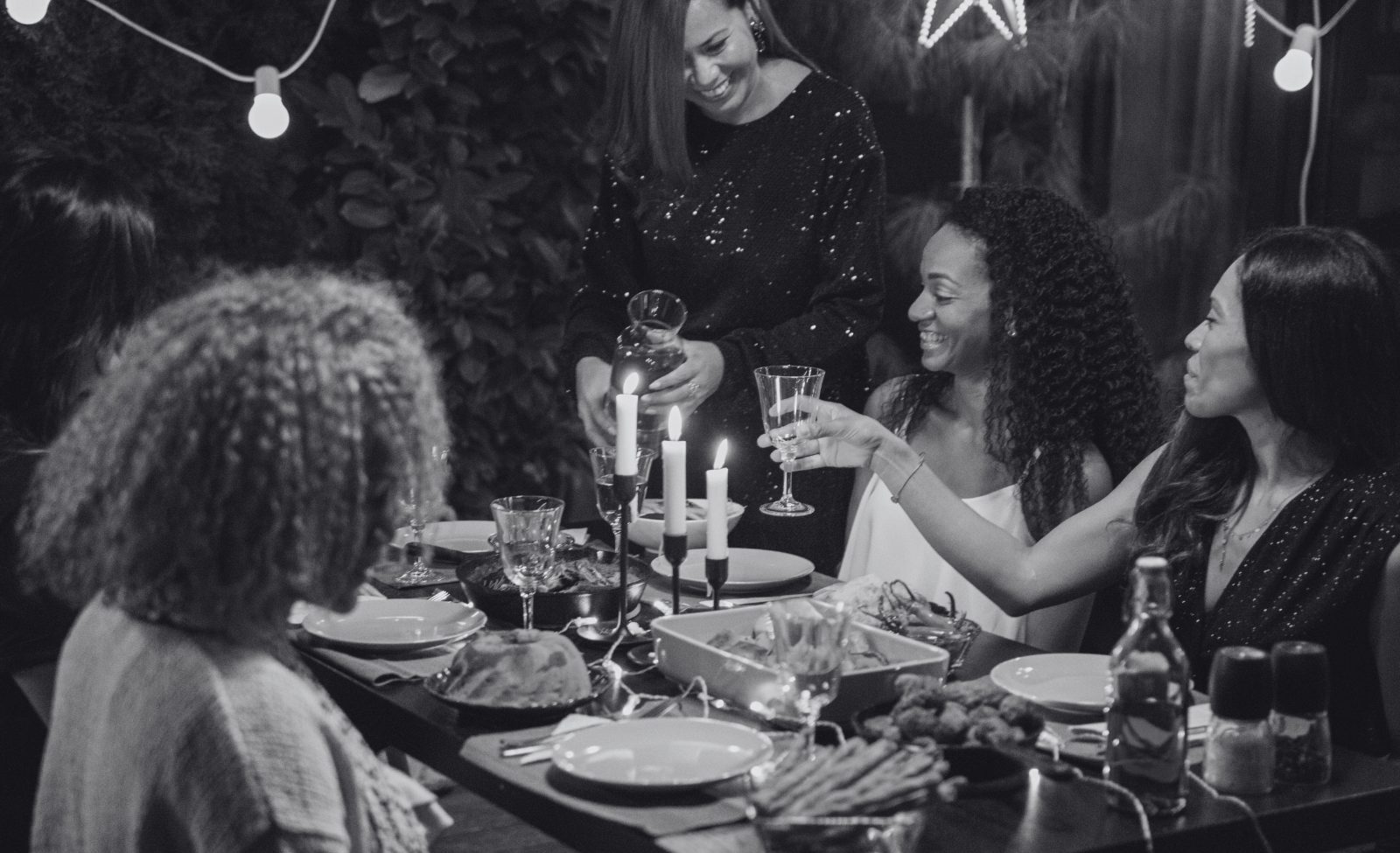Guests laughing around a candlelit dinner table set with glasses and bottles.